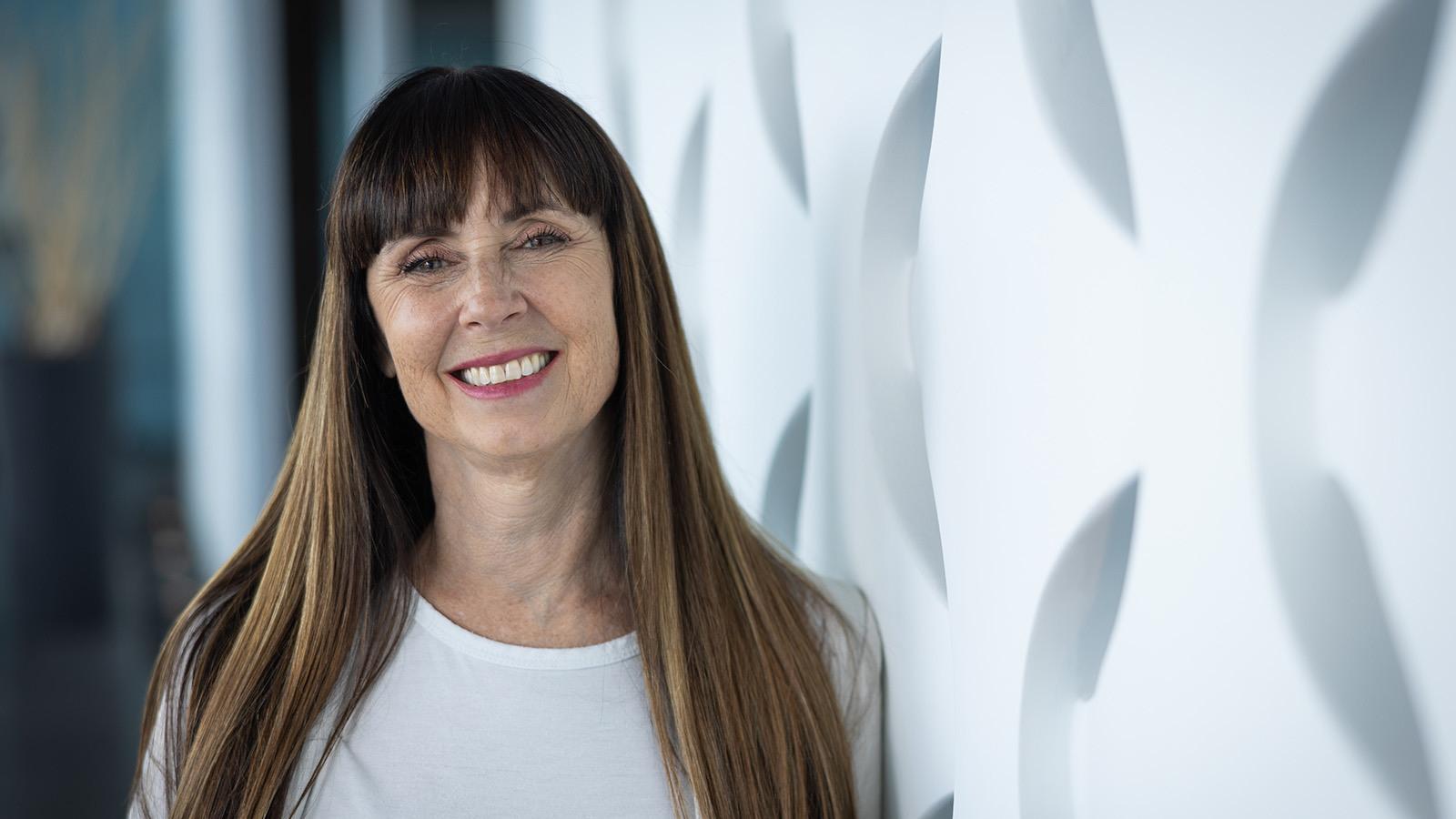 A woman with long brown hair 和 bangs smiling while st和ing against a geometric white backdrop.