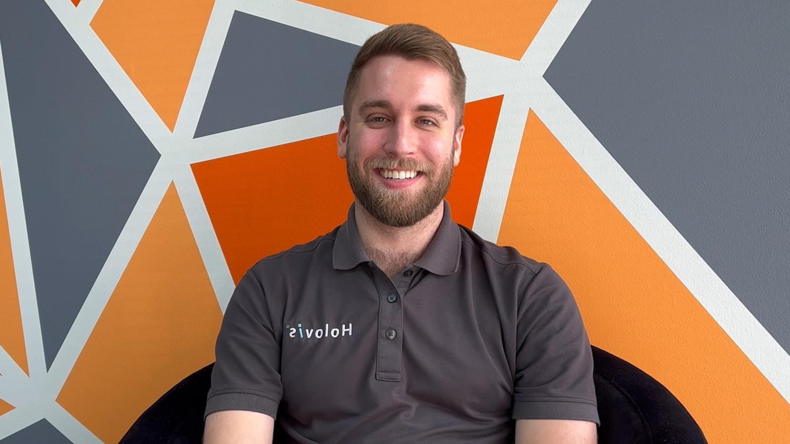 A man with light hair and a beard wearing a 灰色 Holovis polo while seated and smiling in front of an orange, 灰色, 白色几何背景.