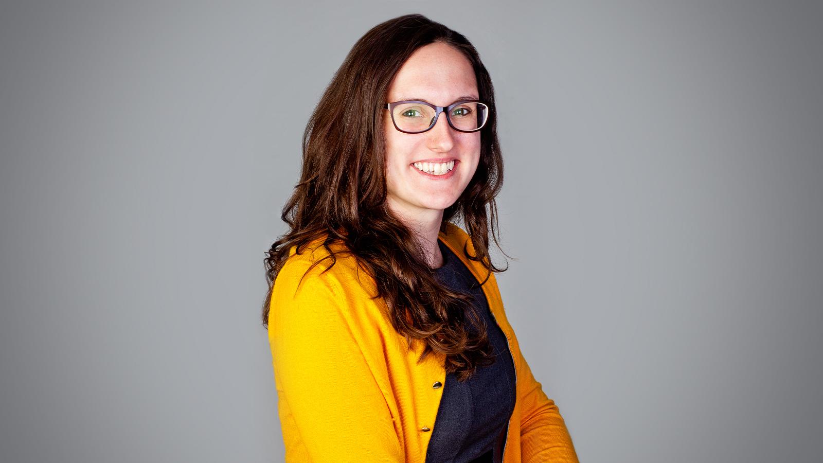 Tasha Rosenbaum smiles at the camera. She has brown hair and is wearing glasses, a yellow cardigan, and a blue sweater.