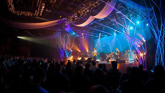 A band plays music in a large, dark auditorium with several bright blue and purple lights pointing at the stage.