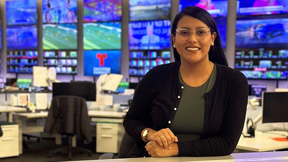 Priscilla sits at a desk in a live news television studio. She is wearing glasses and is smiling.