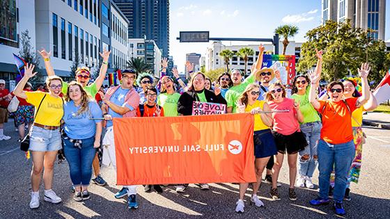 A group of Full Sail faculty, students, and staff posing with a large orange banner that reads “Full Sail University”