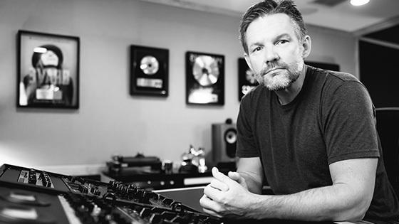 Brad Blackwood sits at an audio console in a studio with framed gold records on the wall behind him.