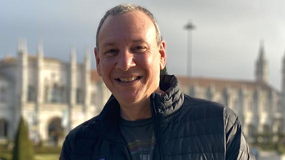 Sean Spuehler stands outdoors in front of a large building. 他面带微笑，穿着一件黑色夹克和一件蓝色t恤.