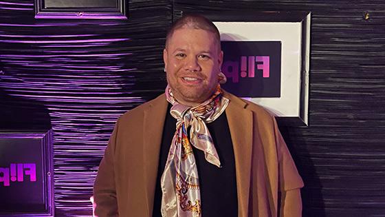 Javi Simons stands in front of a purple wall. 他穿着黑色t恤、牛仔裤、棕色大衣和一条彩色围巾. He is smiling.