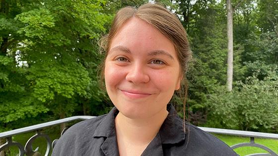 Emmi Litsenberger sits on a porch in front of a grassy lawn and trees. 她面带微笑，穿着一件灰色的纽扣衬衫.