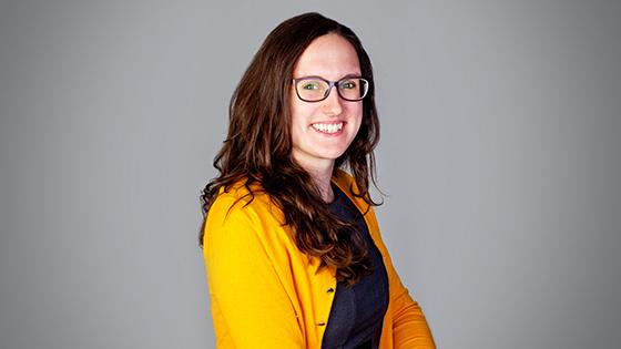 Tasha Rosenbaum smiles at the camera. She has brown hair and is wearing glasses, a yellow cardigan, and a blue sweater.