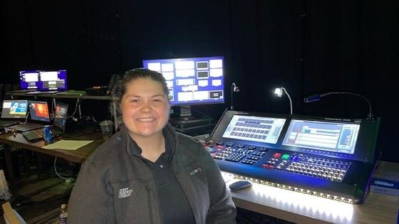 Jean Fuemmeler sits in front of live event video production controllers. She is wearing a gray jacket and smiling.