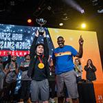 Two people are standing side by side on a stage while one holds a large silver trophy over their head.